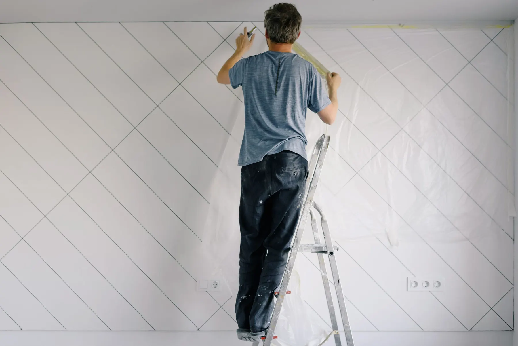 Schilder plakt op een trap de rand langs het plafond af, met folie over de wand gespannen